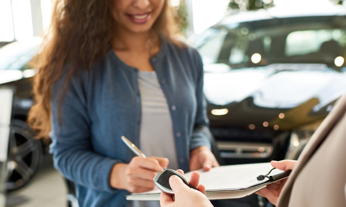 A young woman signing paperwork at a dealership while smiling.