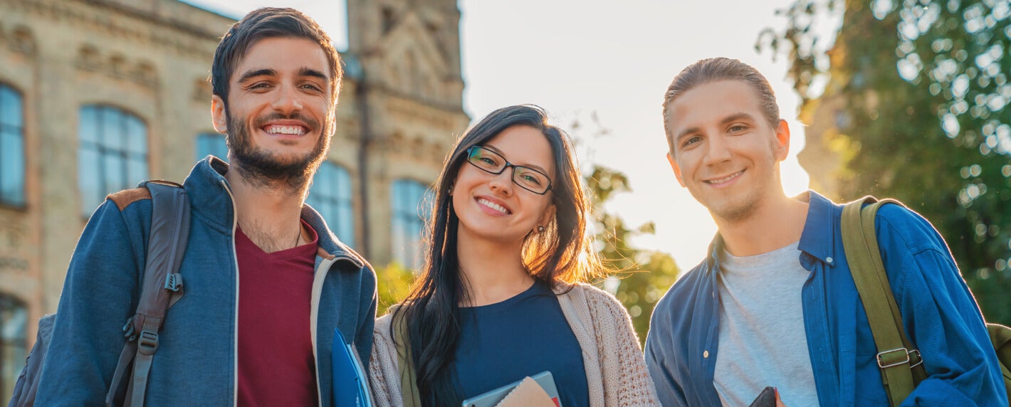 A group of three students standing on a college campus smiling while backlit by the evening sun.