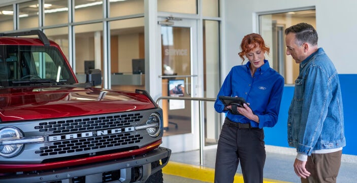 Covert Ford service advisor shows a tablet to a customer beside a red Bronco inside the dealership service center.