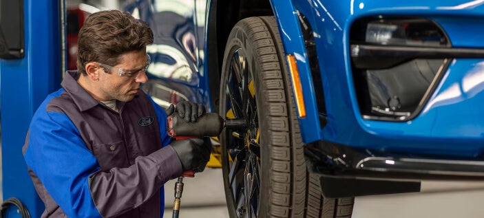 Ford technician removes a wheel from a blue vehicle during service inside a dealership maintenance bay.
