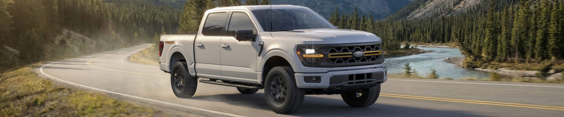 A white Ford F-150 truck drives on a winding road through a scenic mountain landscape with a river and evergreen trees.