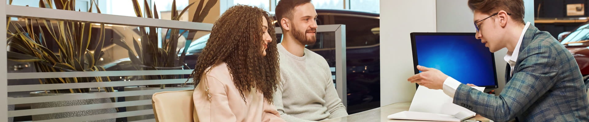 A couple sits at a desk in a car dealership, attentively listening to a salesperson.